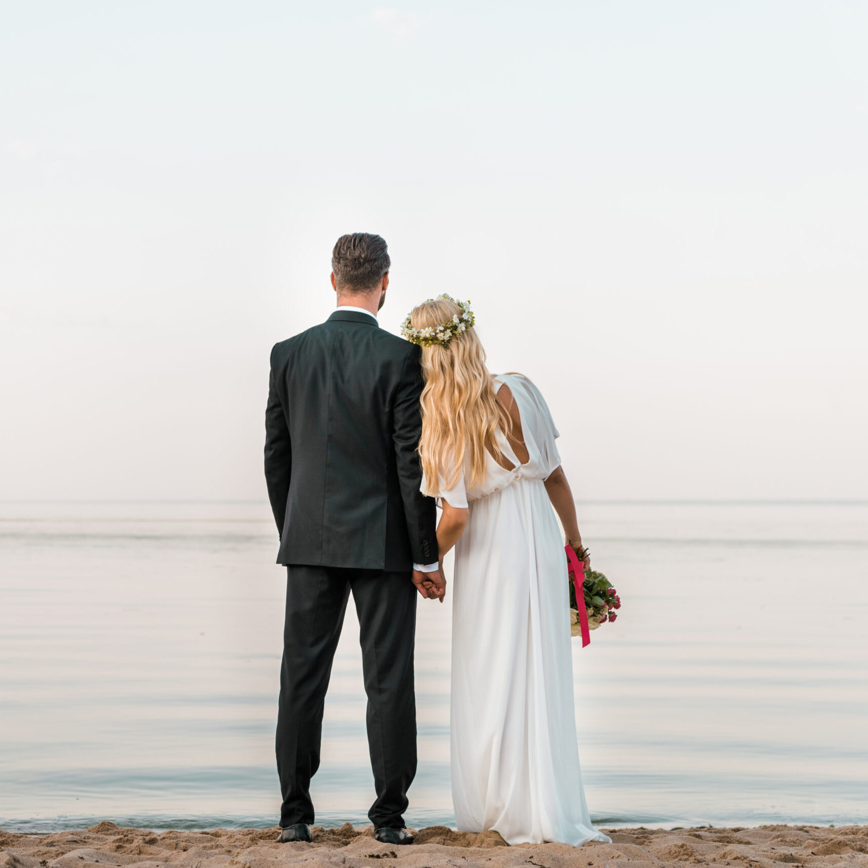 back view of wedding couple standing on beach with wedding bouquet and looking at sea