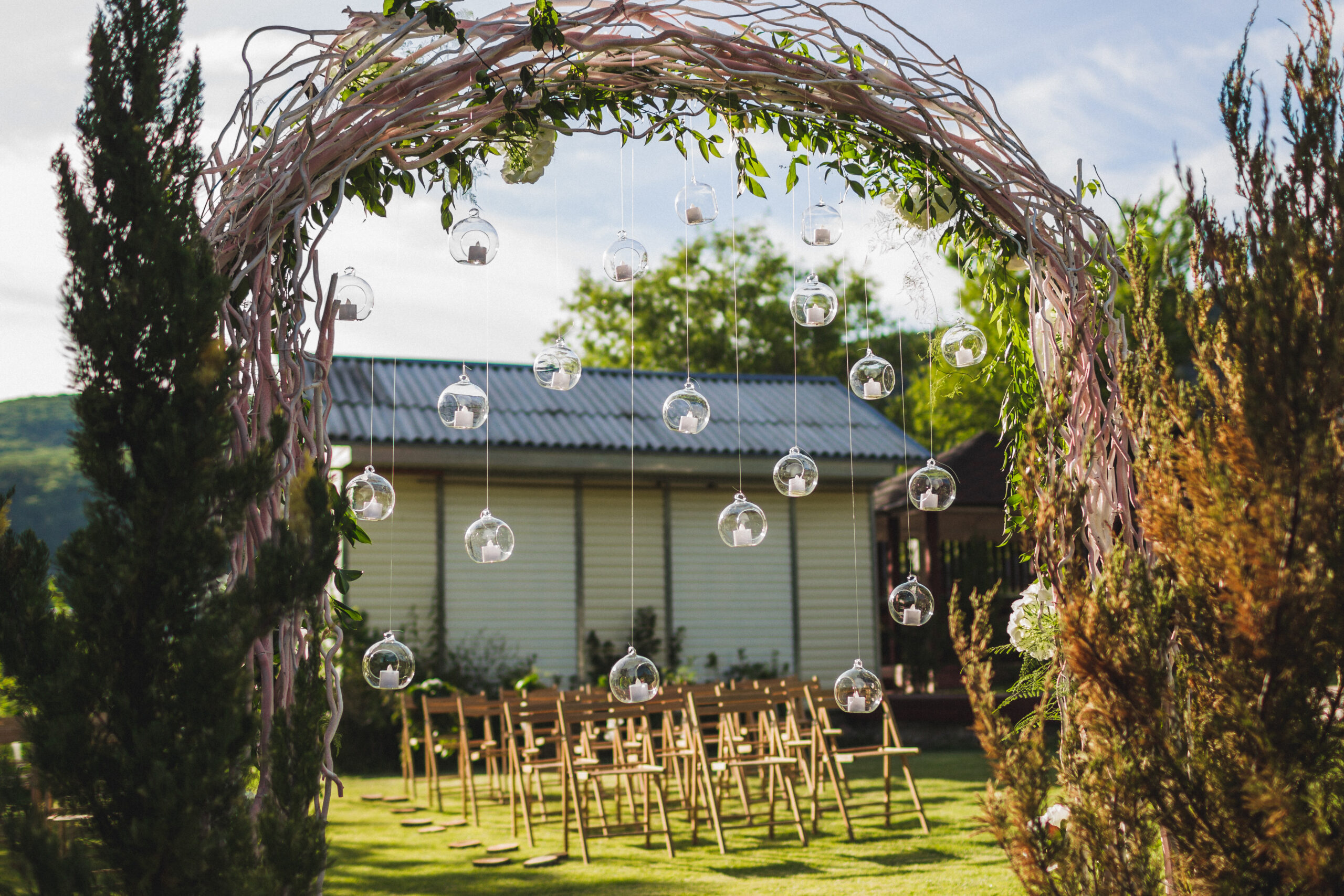 Evening wedding ceremony in garden, arch with white branches, brown wooden chairs and a lot of lights hanging in glass spheres